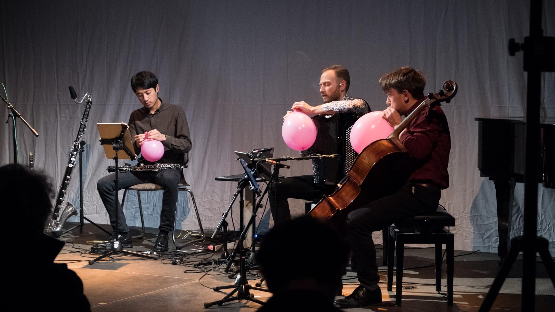 Drei Musiker des Ensembles S201 sitzen auf der Bühne und hantieren mit rosafarbenen Luftballons Drei Musiker des Ensembles S201 sitzen auf der Bühne und hantieren mit rosafarbenen Luftballons