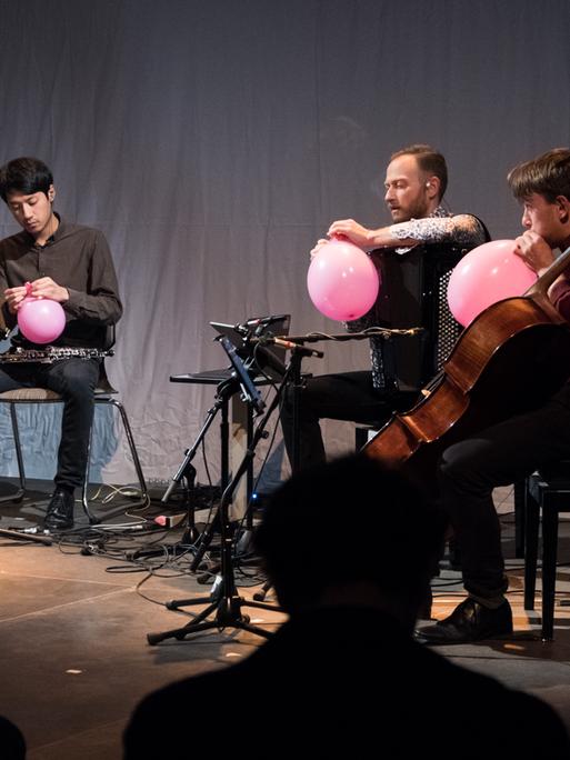 Drei Musiker des Ensembles S201 sitzen auf der Bühne und hantieren mit rosafarbenen Luftballons