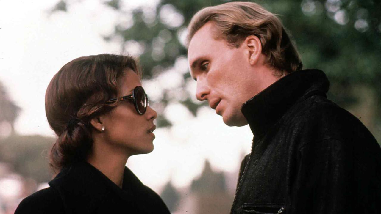 Eine Frau und ein Mann in teurer, dunkler Kleidung stehen sich gegenüber, sie trägt eine dunkle Sonnenbrille. Er lehnt sich mit etwas schrägem Kopf zu ihr hinunter.