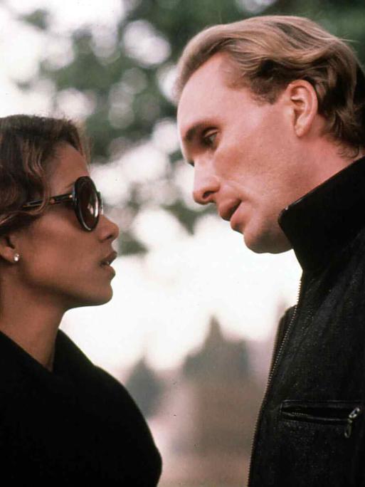 Eine Frau und ein Mann in teurer, dunkler Kleidung stehen sich gegenüber, sie trägt eine dunkle Sonnenbrille. Er lehnt sich mit etwas schrägem Kopf zu ihr hinunter.