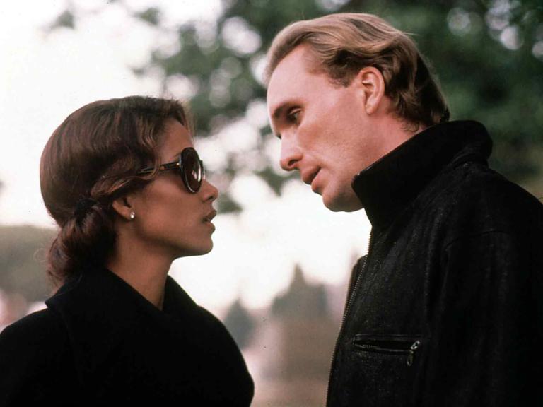 Eine Frau und ein Mann in teurer, dunkler Kleidung stehen sich gegenüber, sie trägt eine dunkle Sonnenbrille. Er lehnt sich mit etwas schrägem Kopf zu ihr hinunter.