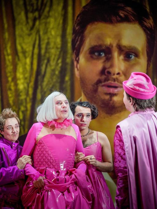Drei Darsteller in rosa und lila Kostümen stehen in Richtung Kamera, einer mit rosa Hut wendet uns den Rücken zu. Im Hintergrund ist ein bärtiges Gesicht auf eine Leinwand projiziert.