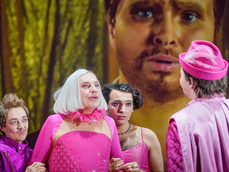 Drei Darsteller in rosa und lila Kostümen stehen in Richtung Kamera, einer mit rosa Hut wendet uns den Rücken zu. Im Hintergrund ist ein bärtiges Gesicht auf eine Leinwand projiziert.