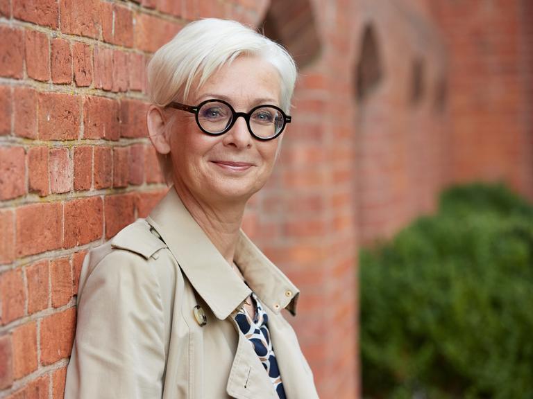 Dörte Hansen, Autorin, bei einem Fototermin. Sie hat kurze graue Haare, rägt einen hellen Trenchcoat und eine prägnante schwarze Brille. Sie steht vor einer Ziegelmauer.