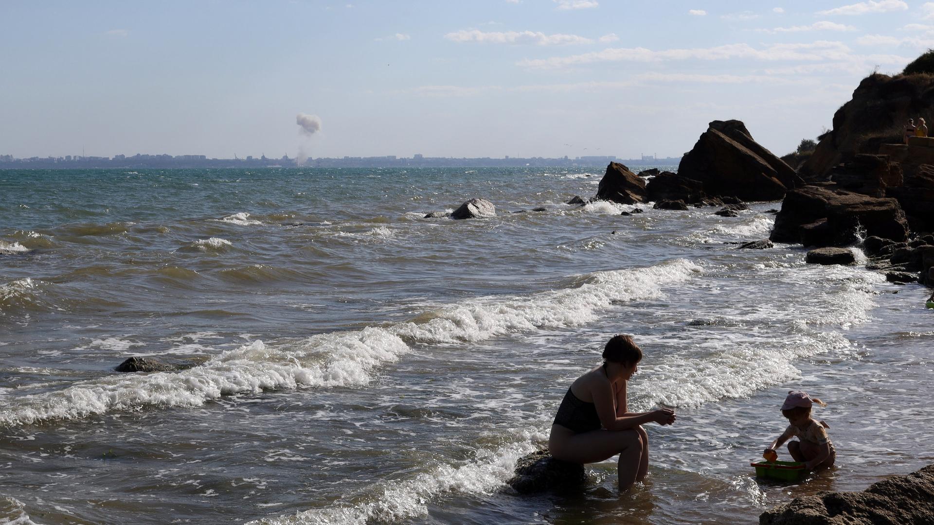 Eine Mutter spielt mit ihrem Kind am Strand von Odessa, während am Horizont eine Bombe im Meer detoniert.
