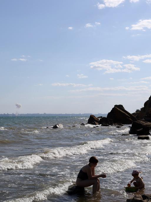 Eine Mutter spielt mit ihrem Kind am Strand von Odessa, während am Horizont eine Bombe im Meer detoniert.