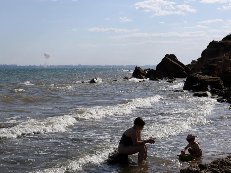 Eine Mutter spielt mit ihrem Kind am Strand von Odessa, während am Horizont eine Bombe im Meer detoniert.