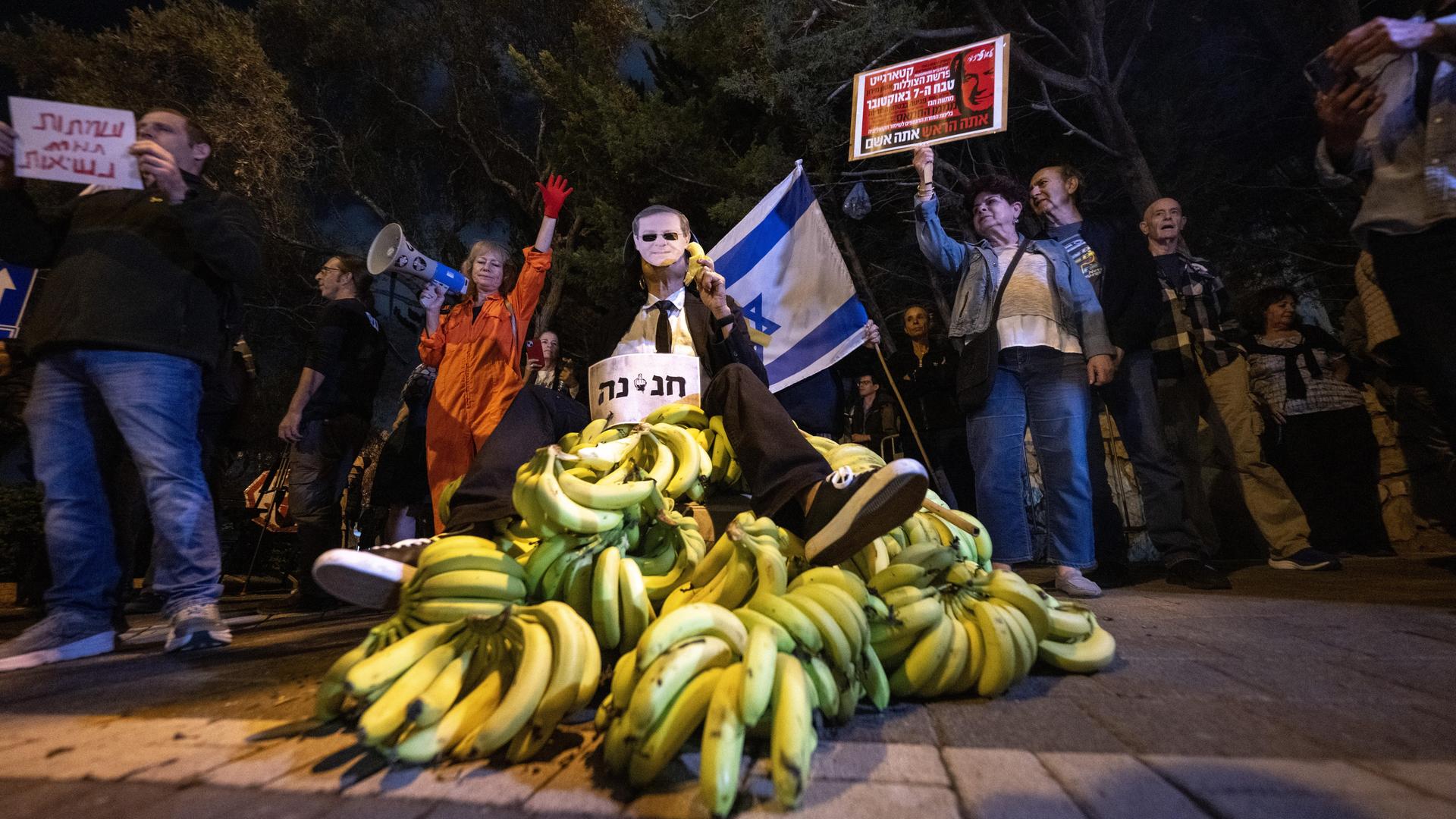 Es ist dunkel. Um einen Haufen Bananen stehen Demonstranten, die Schilder mit heräischer Schrift halten. Eine hält eine Israel-Flagge. Es ist dunkel. Um einen Haufen Bananen stehen Demonstranten, die Schilder mit heräischer Schrift halten. Eine hält eine Israel-Flagge.