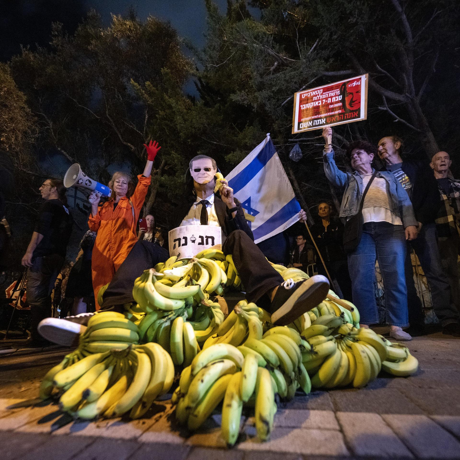 Es ist dunkel. Um einen Haufen Bananen stehen Demonstranten, die Schilder mit heräischer Schrift halten. Eine hält eine Israel-Flagge.