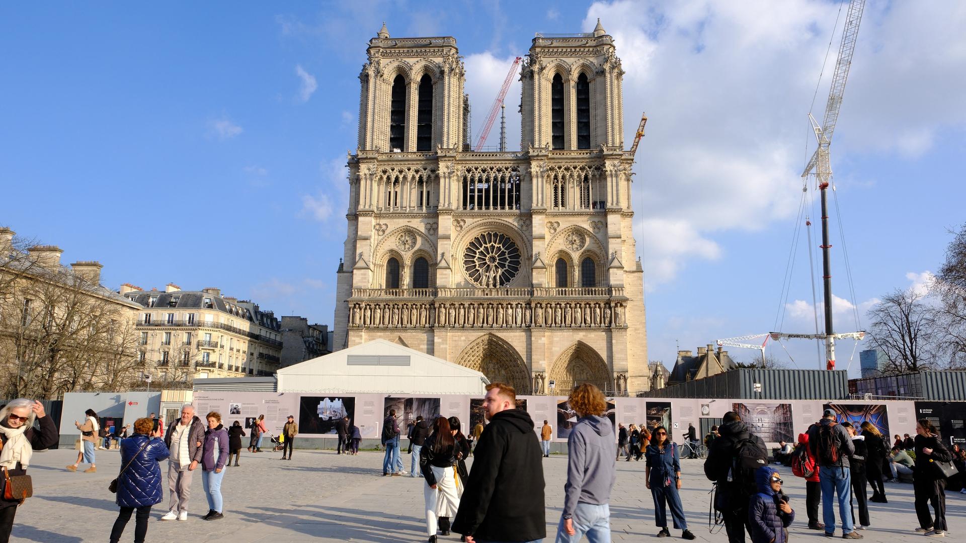 Blick auf die Pariser Kirche Notre Dame: Auf dem Platz im Vordergrund sind herumlaufende Menschen zu sehen, hinter der Kirche ragen zwei Baukräne hervor.