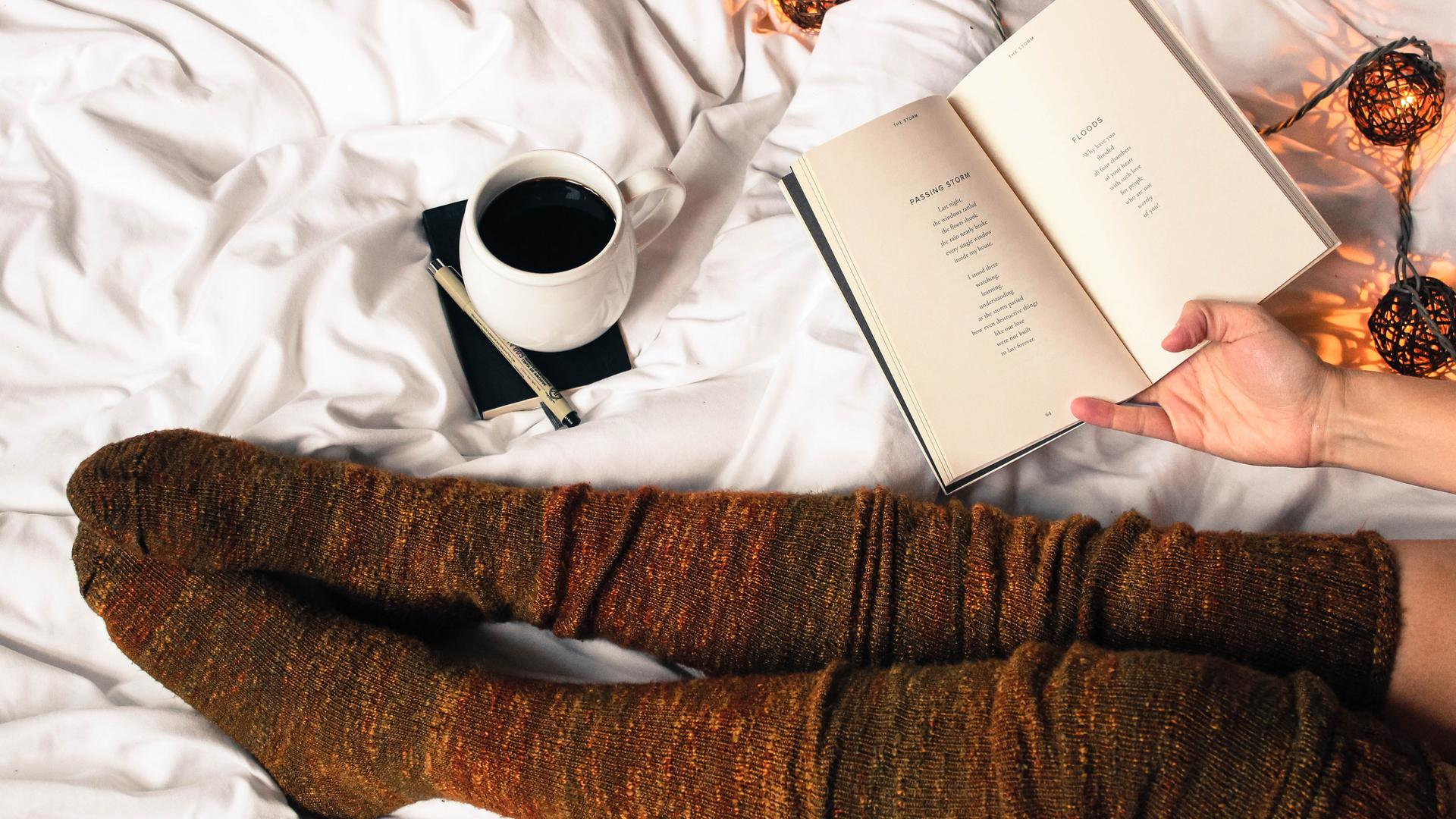 Draufsicht auf ein aufgeschlagenes Buch. Die Beine der Leserin sind sichtbar und in herbstliche, lange Strümpfe gekleidet. Außerdem sieht man eine Tasse Kaffee und Leuchtkugeln. Die Atmosphäre ist herbstlich und behaglich.