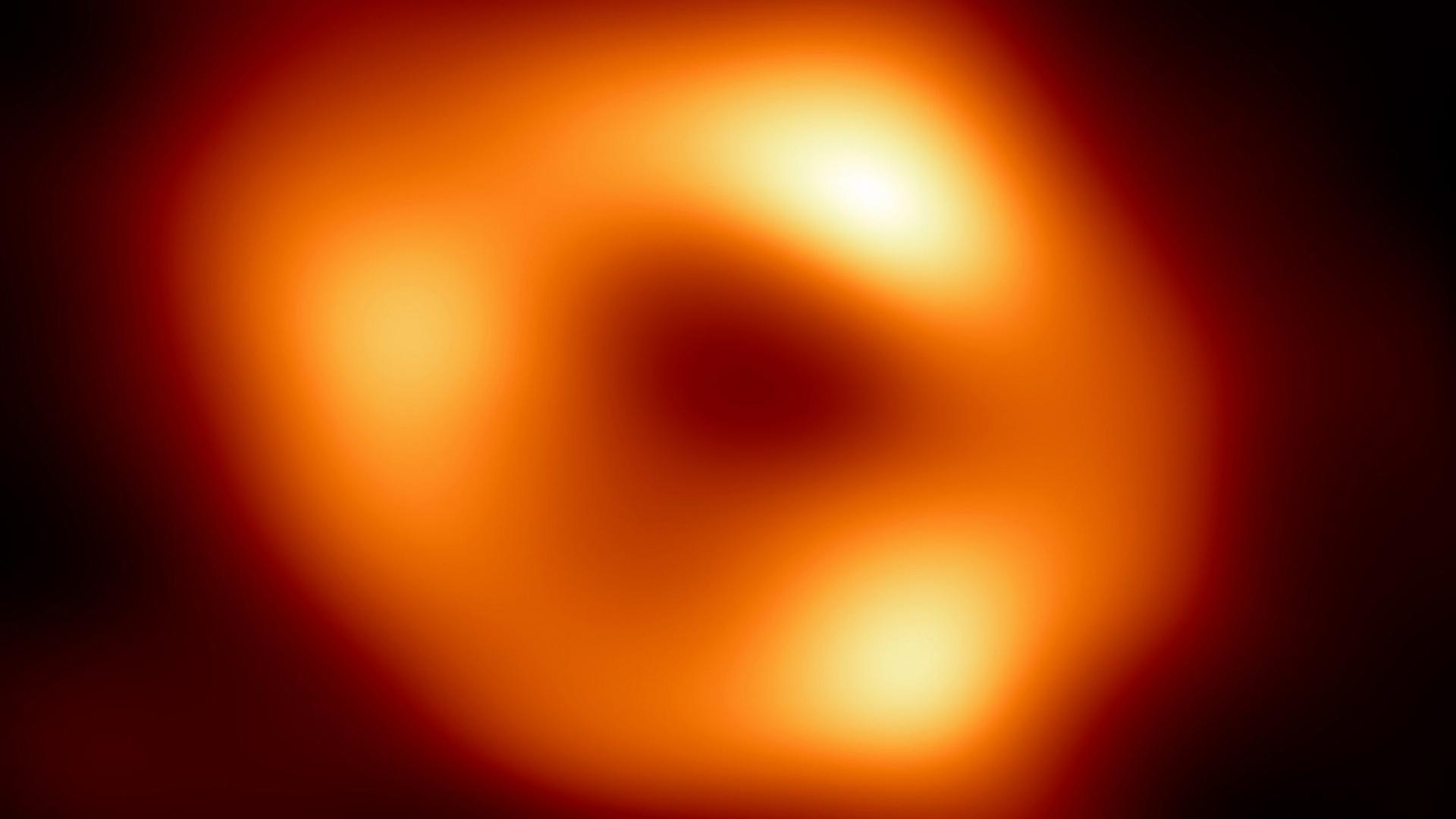 Bild der Event Horizon Telescope Collaboration. Zu sehen ist ein orange-rot-farbener, verschwommener Lichtkreis mit dunklerer Stelle in der Mitte. Außenherum ist es schwarz.