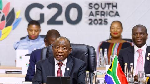 Ramaphosa im dunklen Jackett sitzt am Präsidiumstisch und spricht in ein Mikrofon; dahinter sitzen weitere personen. An einer weißen Wand steht in großen schwarzen Buchstaben "G 20 South Africa 2025". 