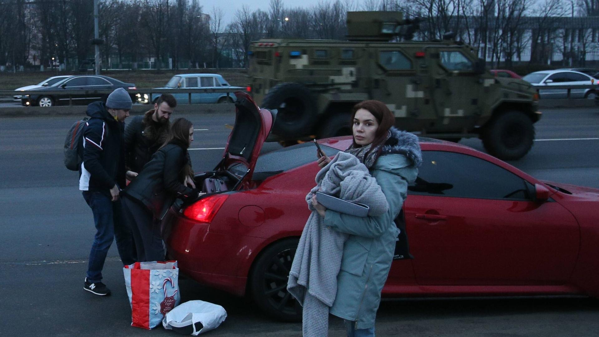 Eine junge Frau auf der Straße mit einem Baby steht vor einem Auto, das Männer mit Gepäck beladen. Dahinter fährt ein Militärfahrzeug.