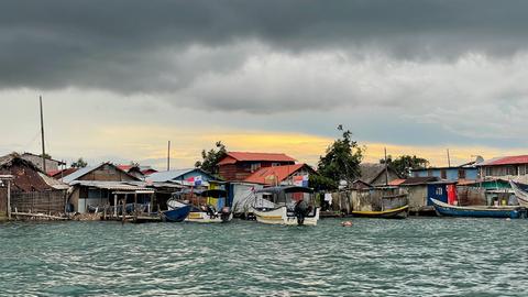 Mitten im Meer liegen dichtgedrängte windschiefe  Holzhütten dicht aneinander. Das Wasser steht fast schon auf der Höhe der Hütten, einige Boote liegen an den Stegen.