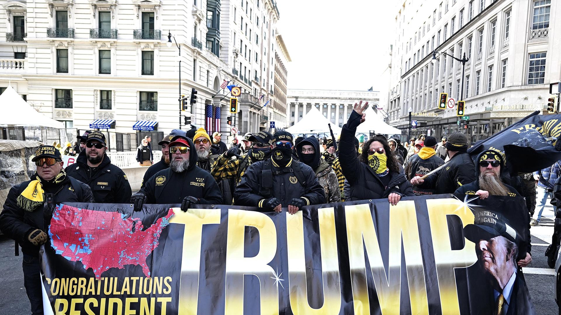 Anhänger der Proud Boys halten ein Banner mit Der Aufschrift "Trump" hoch, sie sind vermummt.