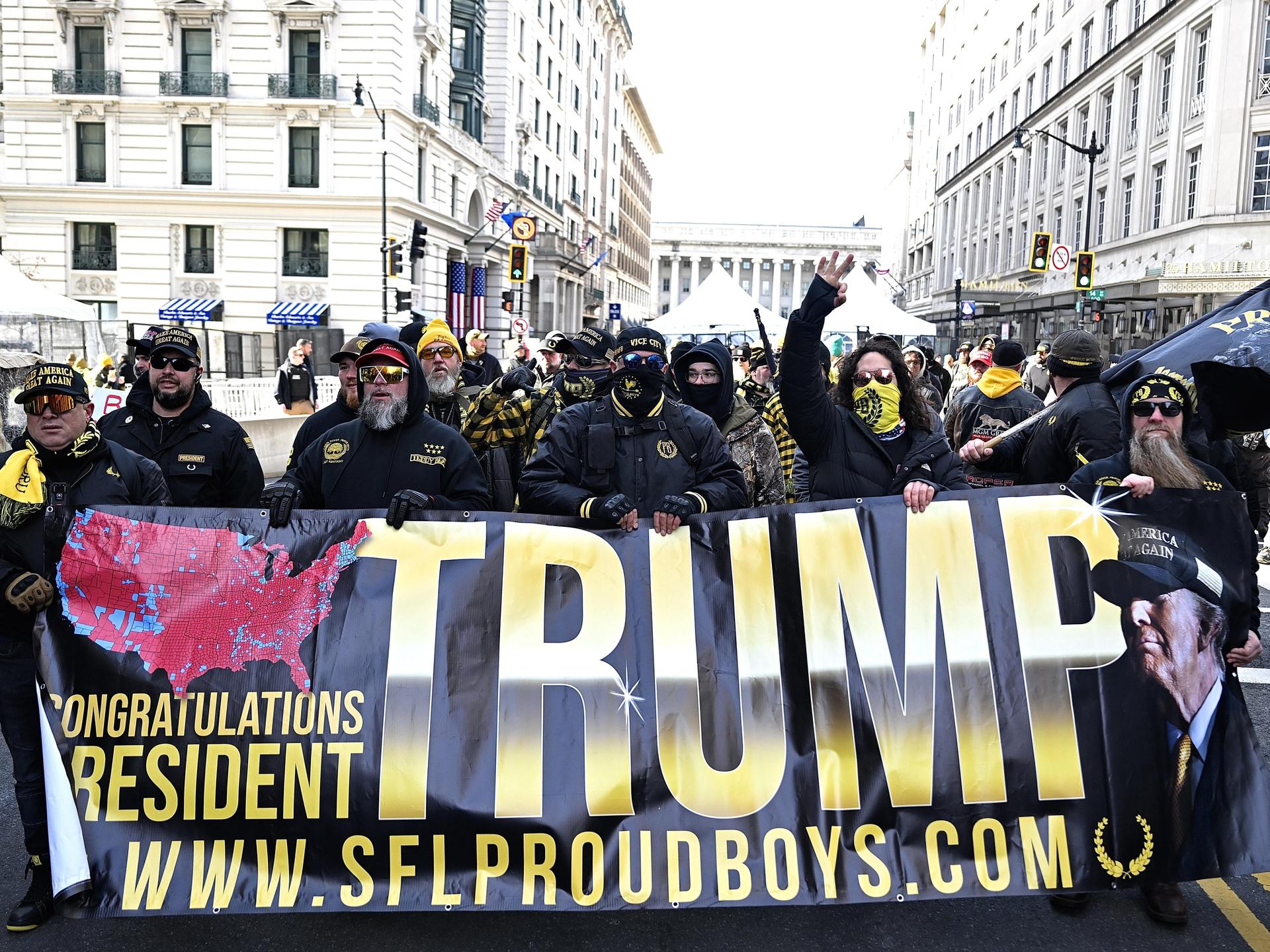 Anhänger der Proud Boys halten ein Banner mit Der Aufschrift "Trump" hoch, sie sind vermummt.