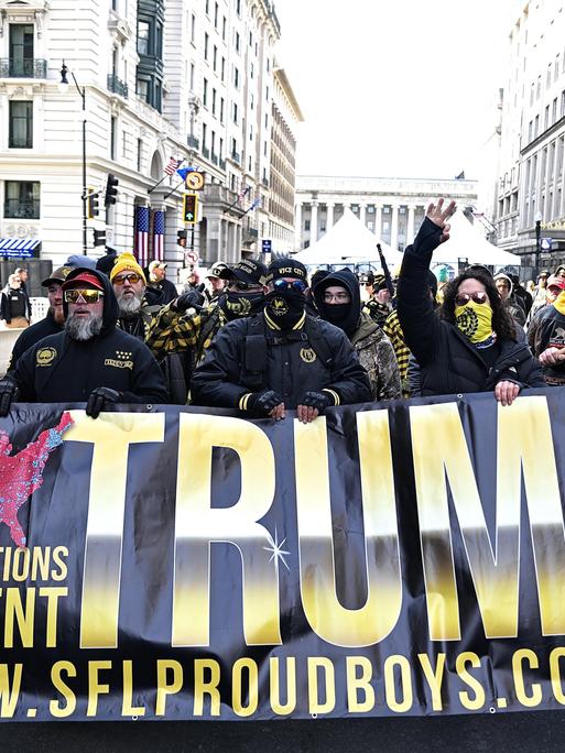 Anhänger der Proud Boys halten ein Banner mit Der Aufschrift "Trump" hoch, sie sind vermummt.