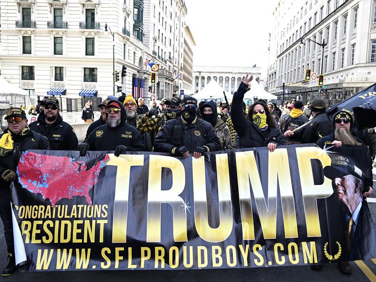Anhänger der Proud Boys halten ein Banner mit Der Aufschrift "Trump" hoch, sie sind vermummt.