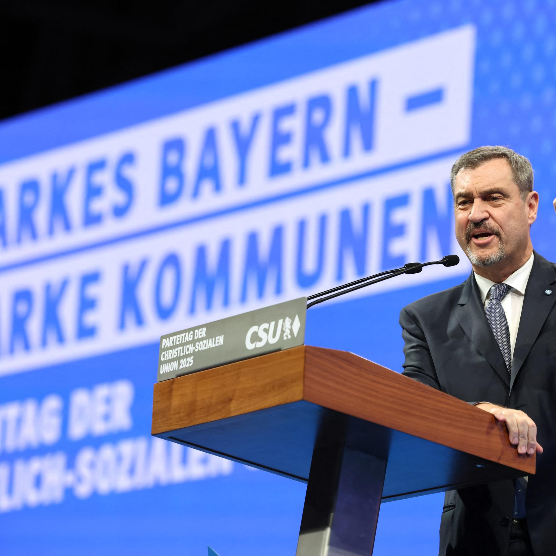 CSU-Chef Söder steht an einem Rednerpult und hebt die linke Hand, um seinen Worten Nachdruck zu verleihen. Im Hintergrund sieht man auf einer großen Tafel das Motto: Starkes Bayern - Starke Kommunen