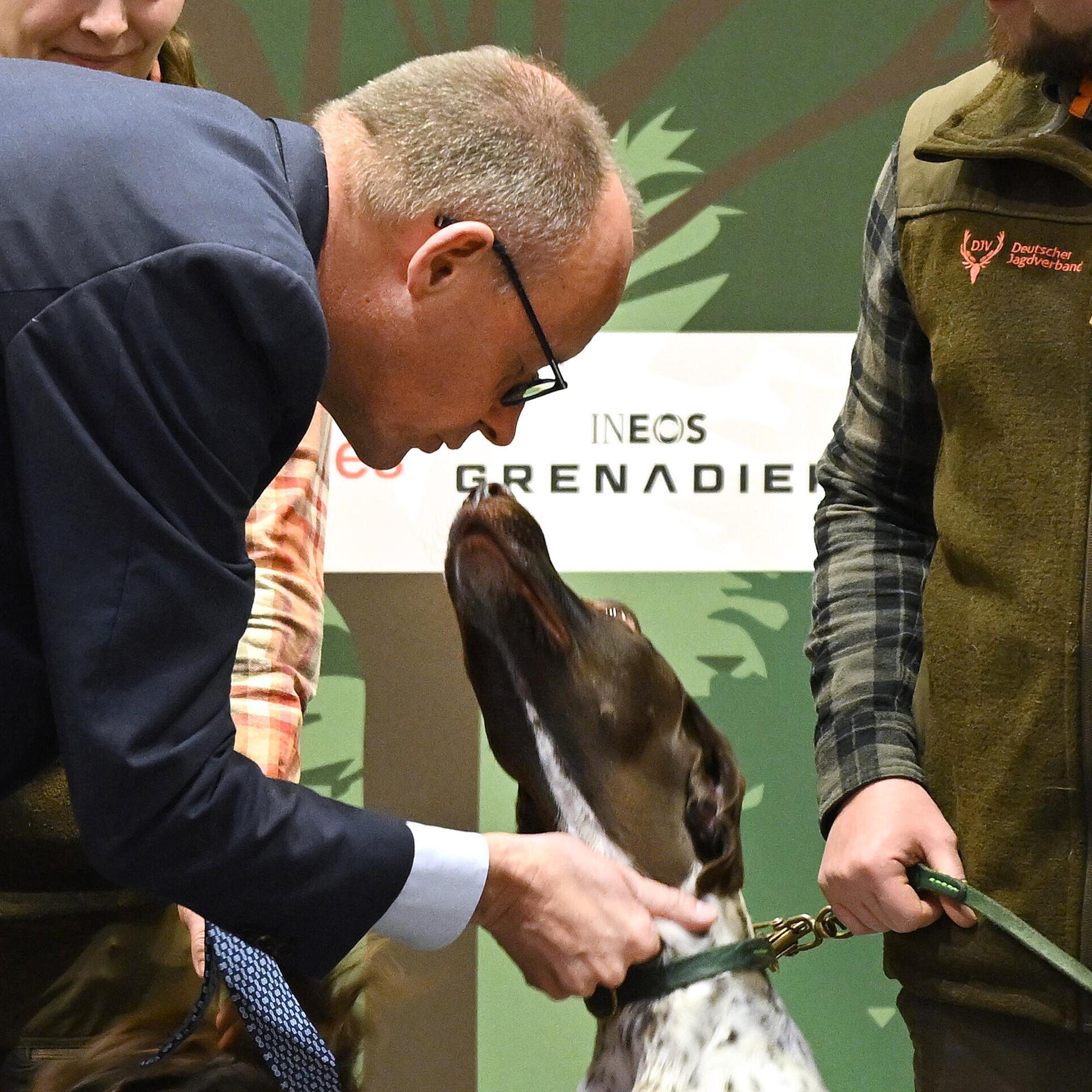 Bundeskanzler Merz trifft bei einem Rundgang auf der Grünen Woche am Stand des Deutschen Jagdverbandes ehrenamtliche Jäger und Jägerinnen mit ihren Hunden. Er beugt sich zu einem Hund herunter und streichelt ihn. 