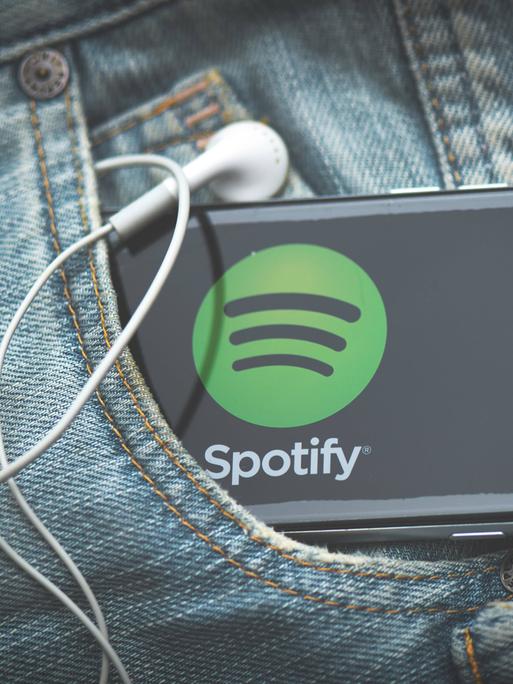 Ein Smartphone mit Spotify-App und Kabelkopfhörern schaut aus einer Jeans-Tasche heraus