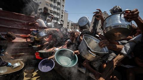 Menschen im Gazastreifen halten an einer Essensausgabe Schüsseln und Töpfe in die Luft. 
