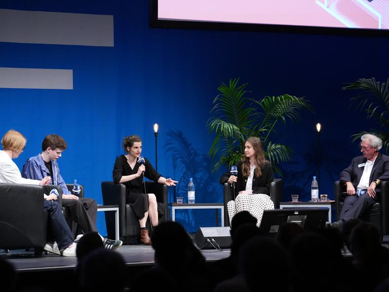 Zu sehen ist die Bühne bei der Europakonferenz im Deutschlandfunk, für die erste Diskussionsrunde sitzen auf Stühlen: der ehemalige luxemburgische Außenminister Jean Asselborn, die Studentin aus Lettland Elizabete Vindule-Mince, Brüssel-Korrespondentin für den Deutschlandfunk Carolin Born und Zivildienstleistender und Aktivist aus Österreich Mati Randow. Ganz links sitzt Moderatorin Katrin Michaelsen.