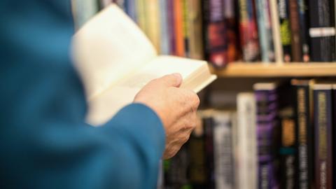 Hände einer Person halten ein Buch. Im Hintergrund ist ein Bücherregal erkennbar.