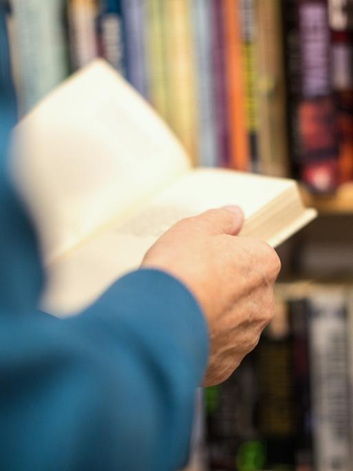 Hände einer Person halten ein Buch. Im Hintergrund ist ein Bücherregal erkennbar.