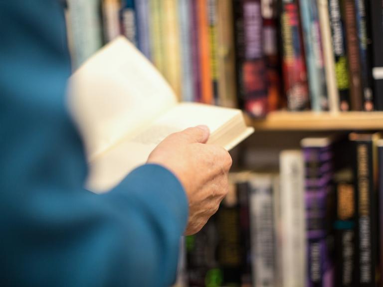 Hände einer Person halten ein Buch. Im Hintergrund ist ein Bücherregal erkennbar.