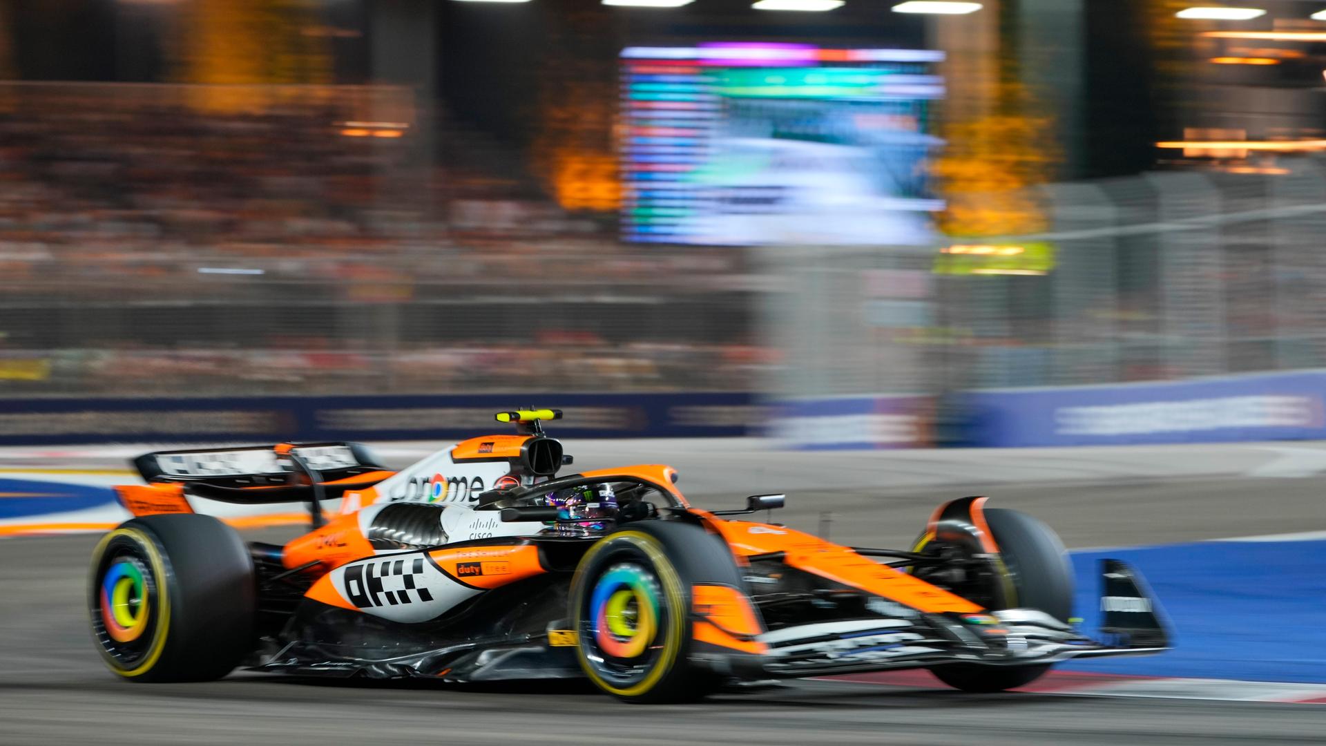 Lando Norris in seinem orange-farbenen McLaren auf der Rennstrecke, dahinter verschwommen eine Leinwand und die Zuschauerränge