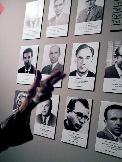 Eine Frau steht vor einer Ausstellungstafel. Darauf sind die Schwarz-Weiß-Bilder von Männern, die an der Entführung von Adolf Eichmann beteiligt waren. Eine Frau steht vor einer Ausstellungstafel. Darauf sind die Schwarz-Weiß-Bilder von Männern, die an der Entführung von Adolf Eichmann beteiligt waren.