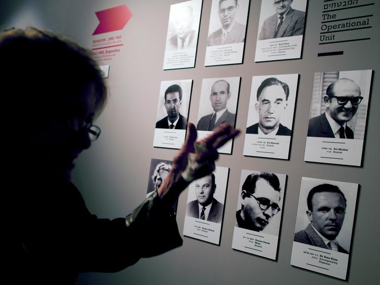 Eine Frau steht vor einer Ausstellungstafel. Darauf sind die Schwarz-Weiß-Bilder von Männern, die an der Entführung von Adolf Eichmann beteiligt waren. Eine Frau steht vor einer Ausstellungstafel. Darauf sind die Schwarz-Weiß-Bilder von Männern, die an der Entführung von Adolf Eichmann beteiligt waren.