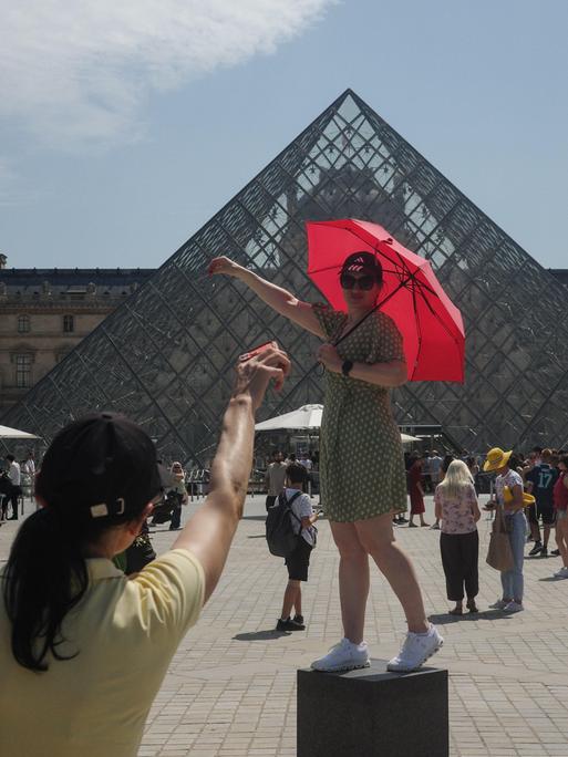 Touristen posieren vor der gläsernen Pyramide des Louvre mit einem Regenschirm als Sonnenschutz