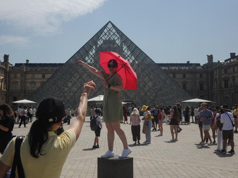 Touristen posieren vor der gläsernen Pyramide des Louvre mit einem Regenschirm als Sonnenschutz