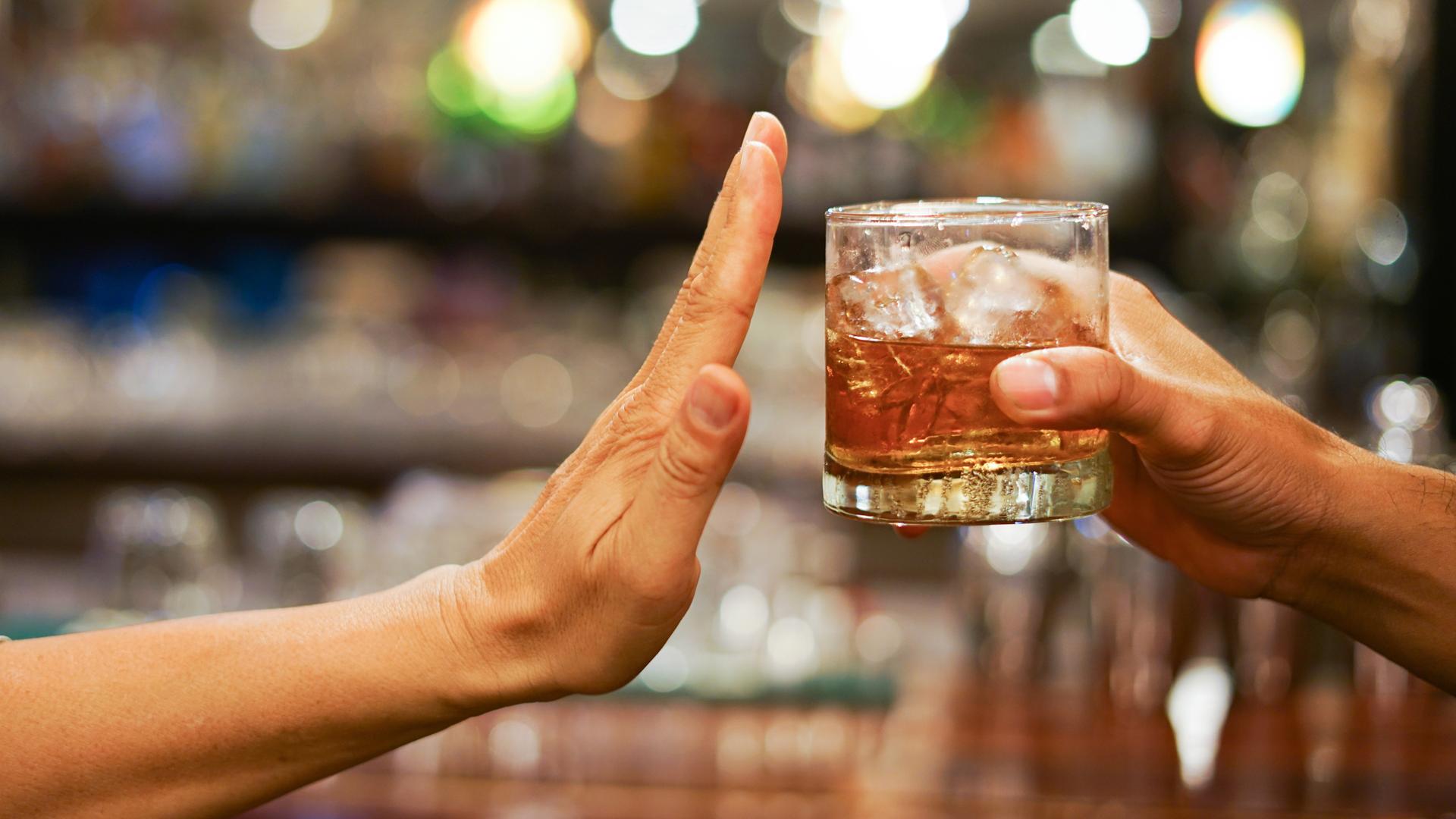 Eine Hand reicht ein Glas mit einer alkoholähnlichen Flüssigkeit von rechts ins Bild. Eine andere bedeutet von der linken Seite "Stop".