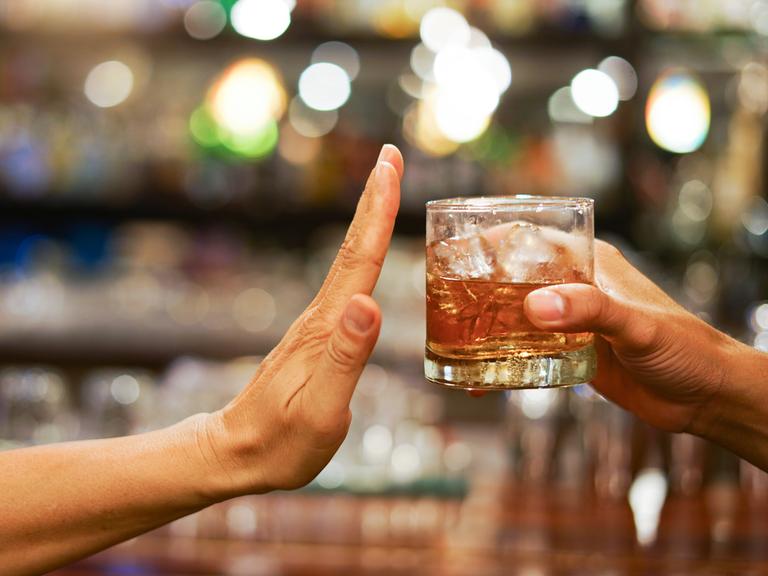 Eine Hand reicht ein Glas mit einer alkoholähnlichen Flüssigkeit von rechts ins Bild. Eine andere bedeutet von der linken Seite "Stop".