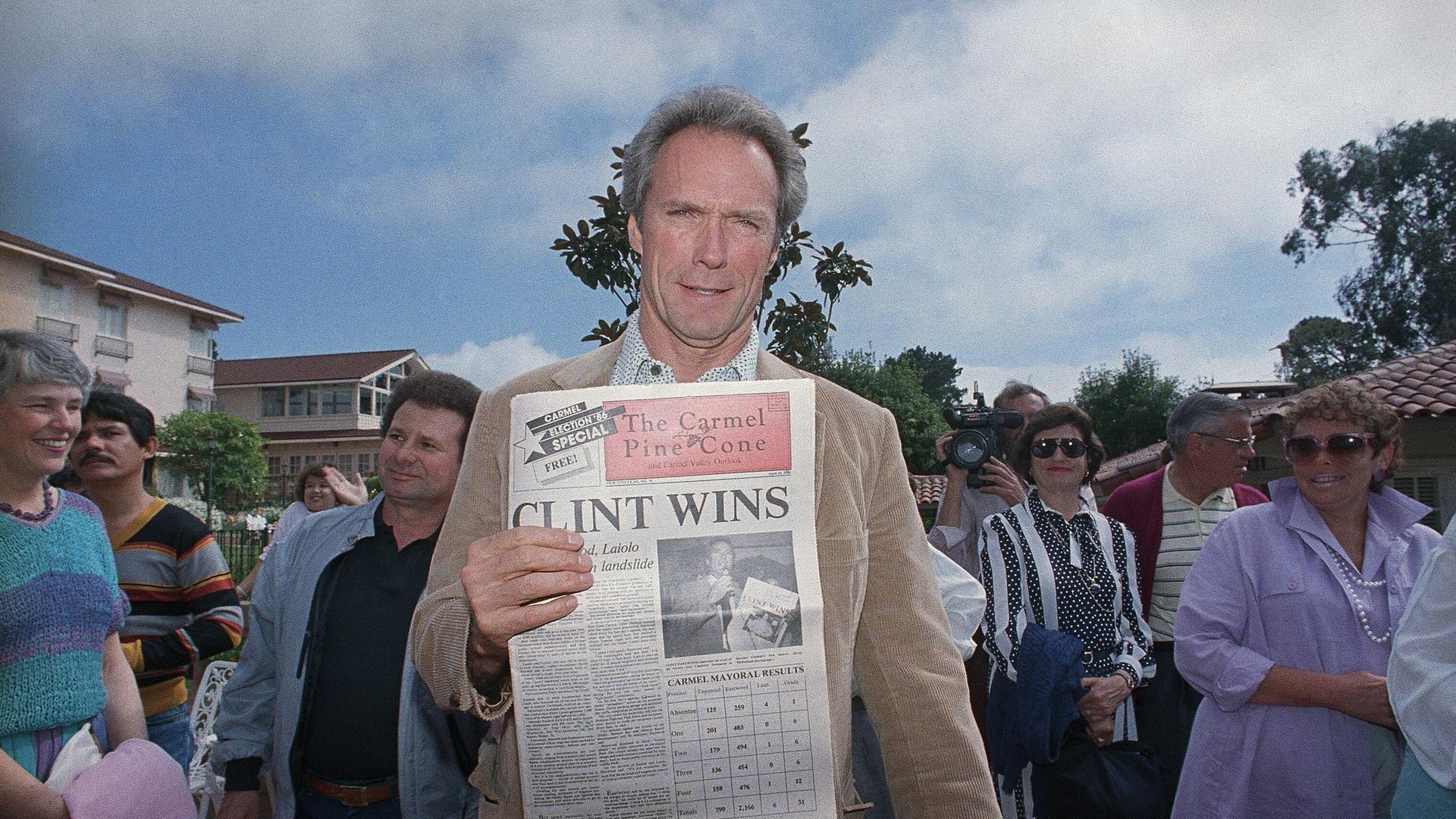 Clint Eastwood steht in Carmel-by-the-Sea zwischen Menschen und hält eine Ausgabe der Zeitung "The Carmel Pine Cone" in die Kamera, der Titel: "CLINT WINS".
