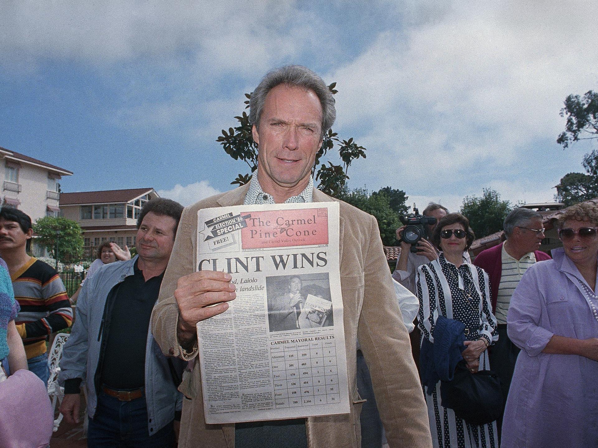 Clint Eastwood steht in Carmel-by-the-Sea zwischen Menschen und hält eine Ausgabe der Zeitung "The Carmel Pine Cone" in die Kamera, der Titel: "CLINT WINS".

