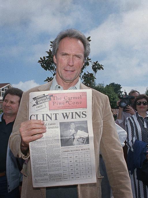 Clint Eastwood steht in Carmel-by-the-Sea zwischen Menschen und hält eine Ausgabe der Zeitung "The Carmel Pine Cone" in die Kamera, der Titel: "CLINT WINS".
