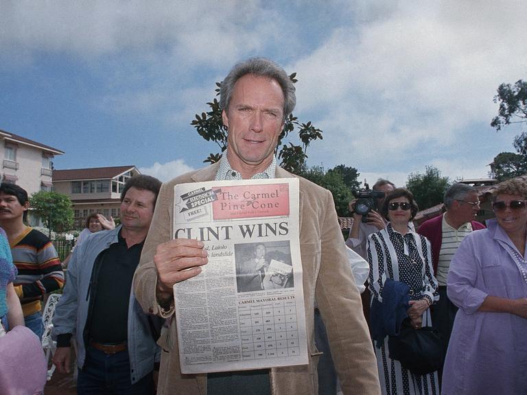 Clint Eastwood steht in Carmel-by-the-Sea zwischen Menschen und hält eine Ausgabe der Zeitung "The Carmel Pine Cone" in die Kamera, der Titel: "CLINT WINS".
