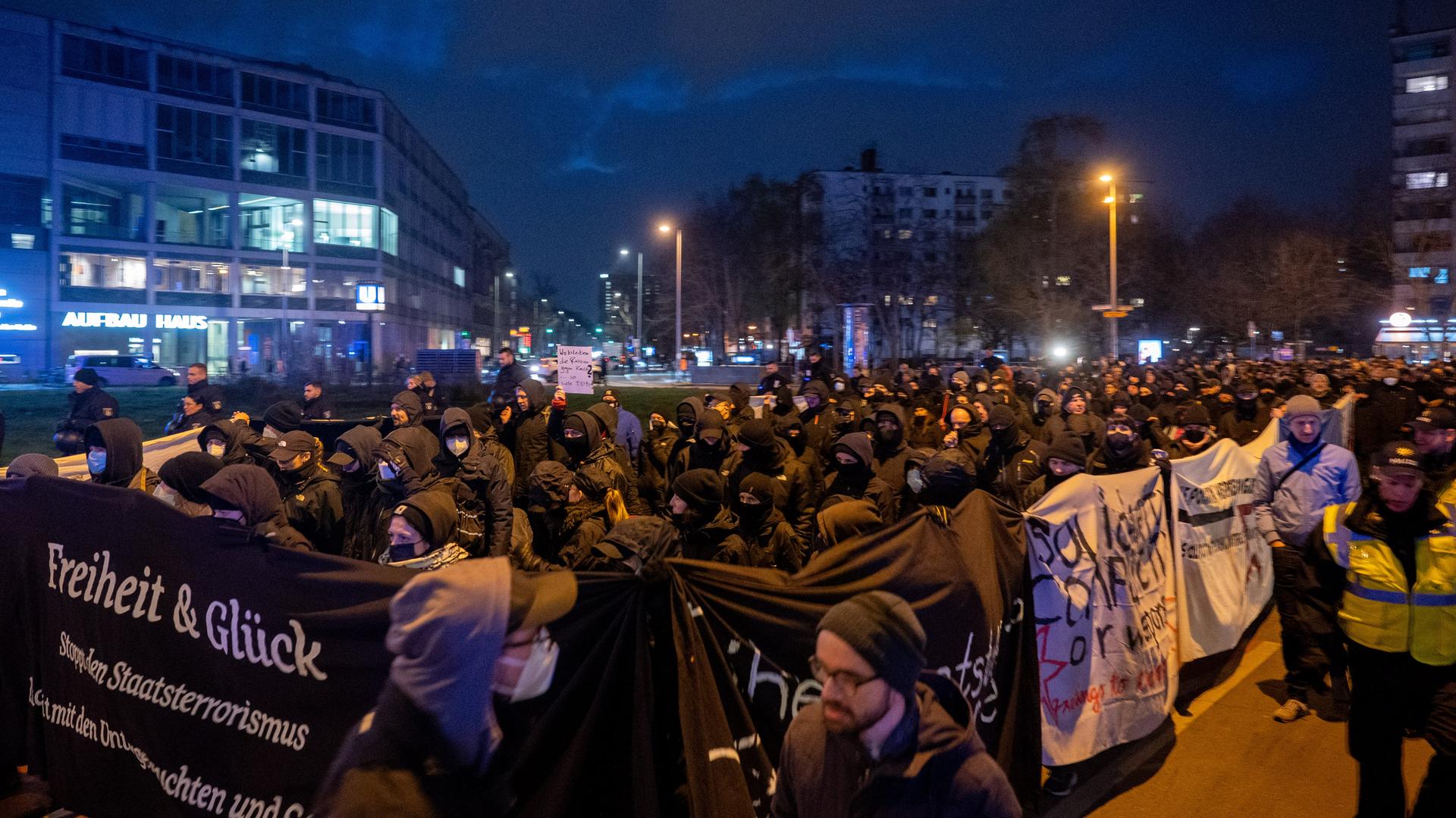Schwarz vermummte Menschen laufen in einem Demonstrationszug mit. Einige tragen Transparente. Auf einem Transparent steht "Freiheit und Glück".