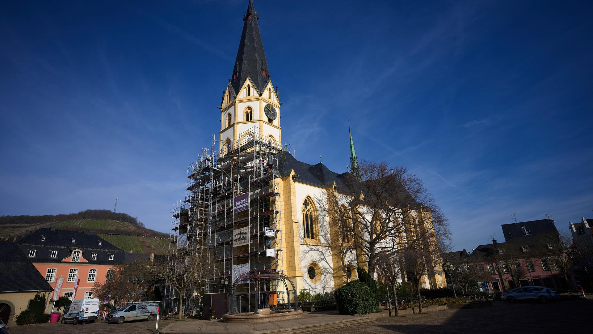 Blick von außen auf die ockerfarben-weiß angestrichene Kirche, an deren Frontseite ein Baugerüst steht. Daneben Häuser, darüber ein blauer Himmel. 
