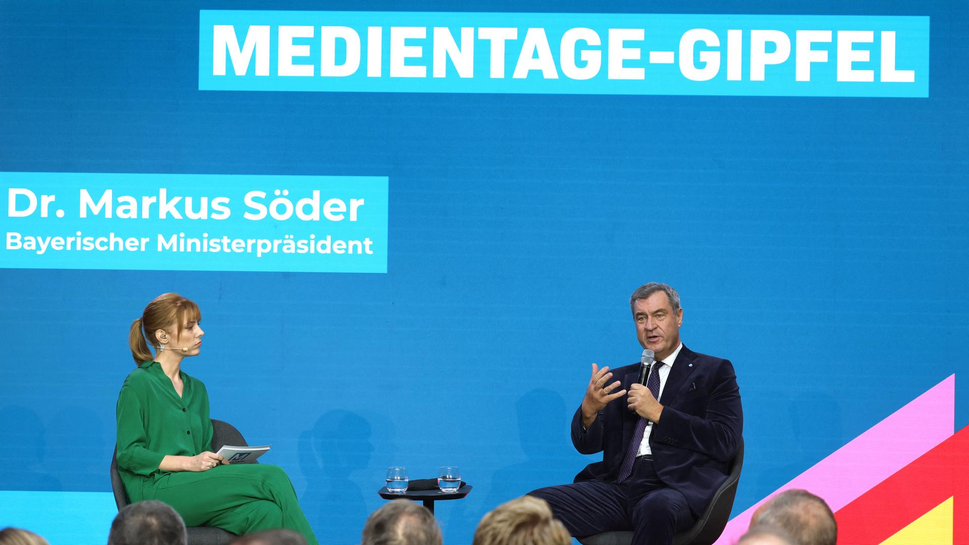 Bayerns Ministerpräsident Markus Söder sitzt bei der Eröffnung der 40. Medientage München auf dem Podium. Neben ihmn sietzt eine in grün gekleidete Frau. 