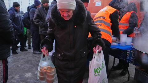 Eine alte Frau trägt Tüten mit Essen davon. Im Hintergrund Männer mit orangen Westen einer Hilfsorganisation, die warme Speisen ausgibt. 