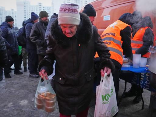 Eine alte Frau trägt Tüten mit Essen davon. Im Hintergrund Männer mit orangen Westen einer Hilfsorganisation, die warme Speisen ausgibt. Eine alte Frau trägt Tüten mit Essen davon. Im Hintergrund Männer mit orangen Westen einer Hilfsorganisation, die warme Speisen ausgibt.