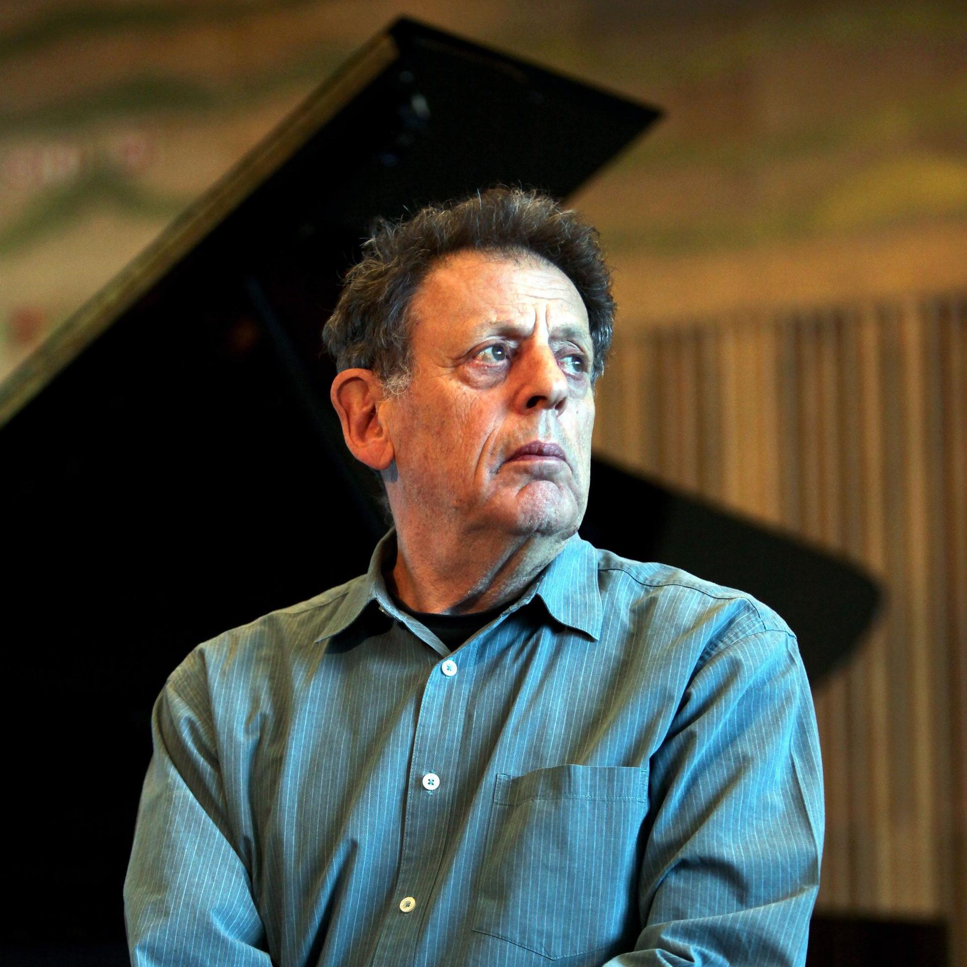 Philip Glass sitzt vor einem aufgeklappten Flügel und schaut nach rechts an der Kamera vorbei.