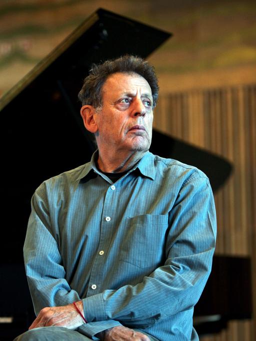 Philip Glass sitzt vor einem aufgeklappten Flügel und schaut nach rechts an der Kamera vorbei.