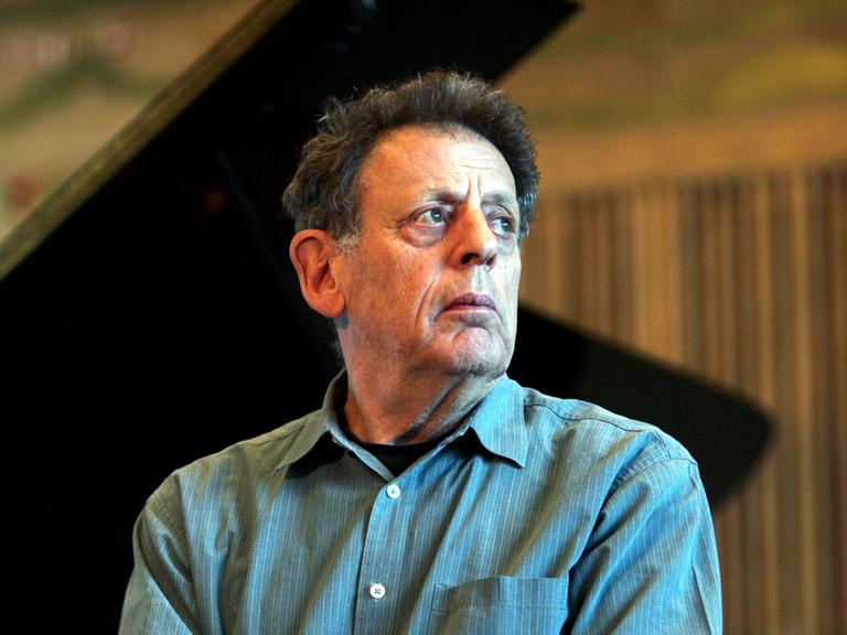 Philip Glass sitzt vor einem aufgeklappten Flügel und schaut nach rechts an der Kamera vorbei.